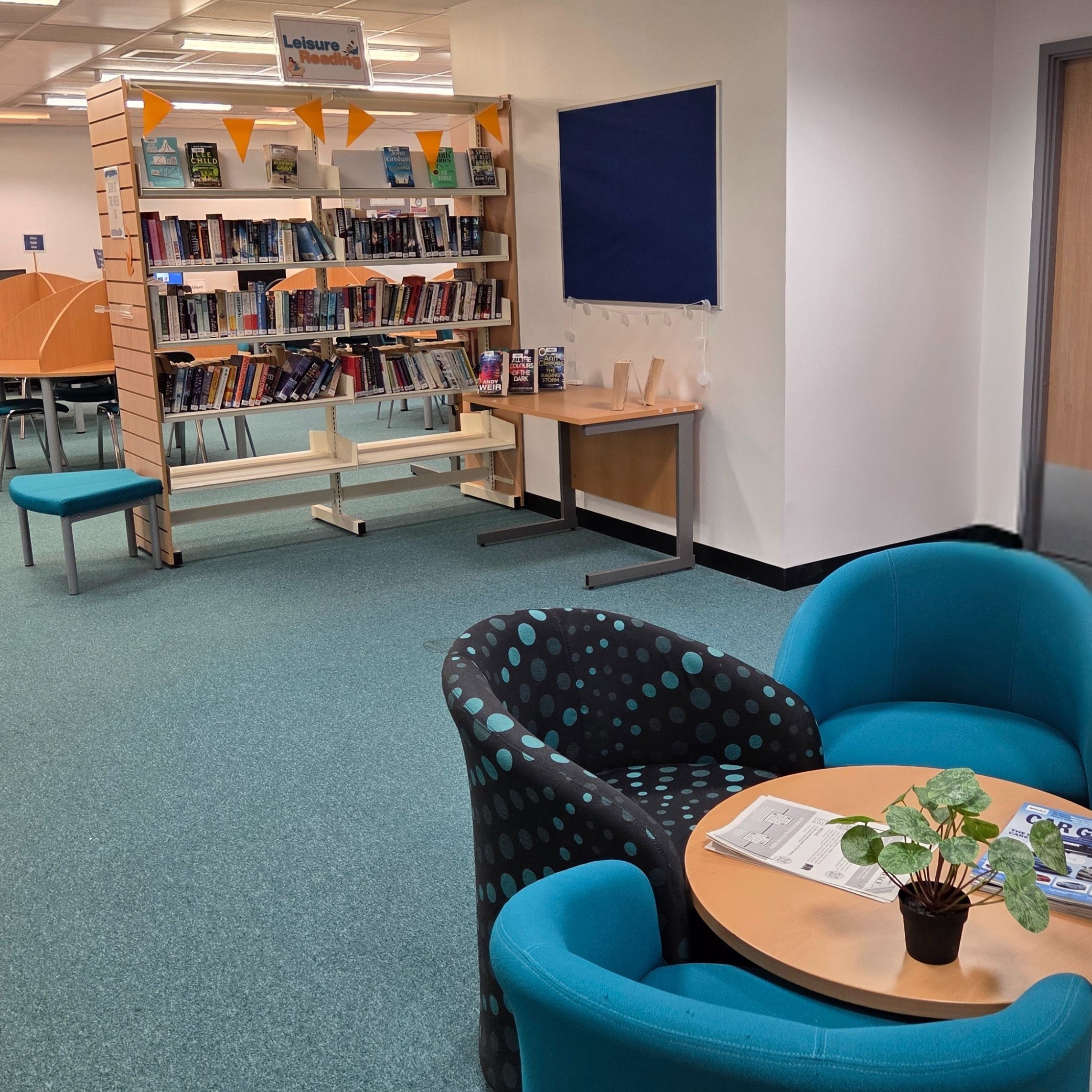 Book shelves filled with books, with study desks to the left and behind it. In front of the book shelf is a table displaying 5 books and  in the foreground is a round coffee table with some chairs