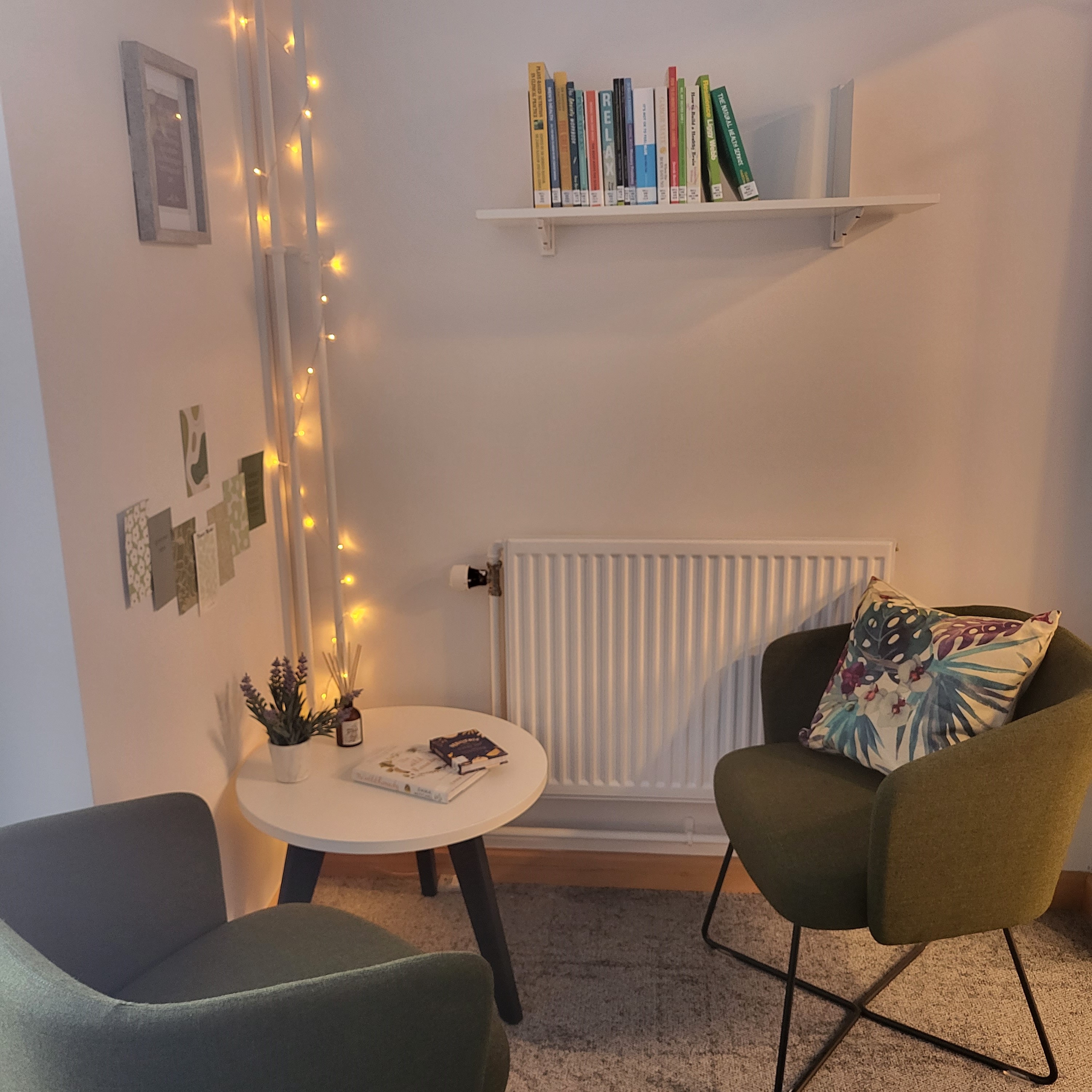 Image shows 2 chairs in the foreground, with a small round table in the corner, there is a shelf with books on it on the wall facing. On the left wall there are some pictures and in the corner going from top to bottom is a string of lights