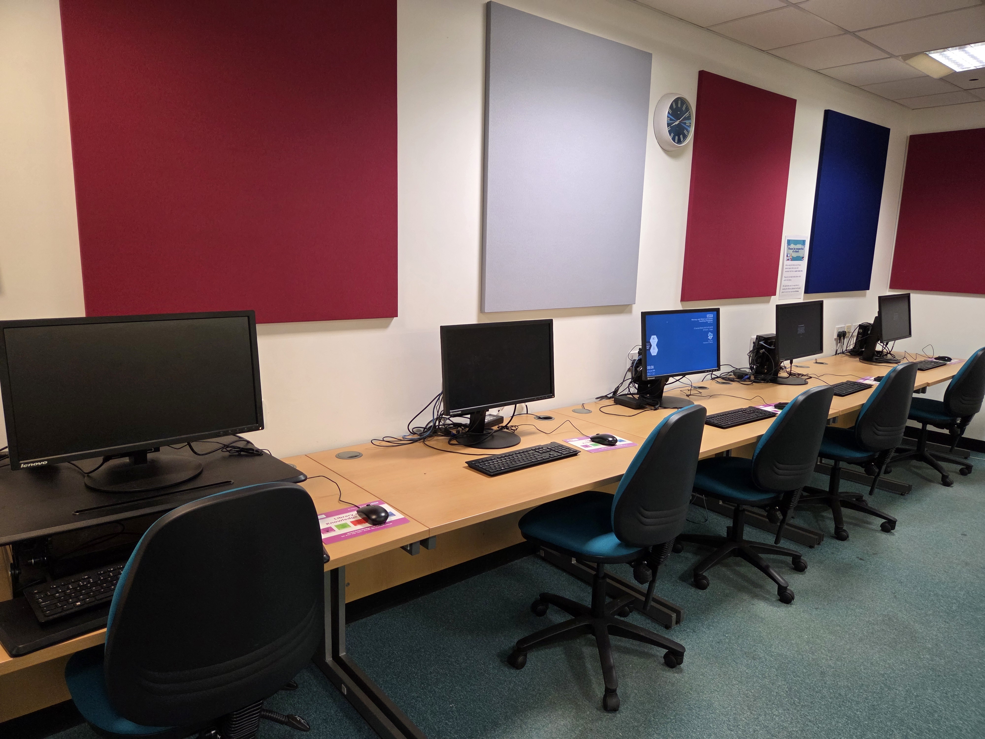 On the wall is a row of 4 panels in a landscape view with a clock in between the centre two. Below is a row of desks with 5 computers on them and 5 chairs under the desks.