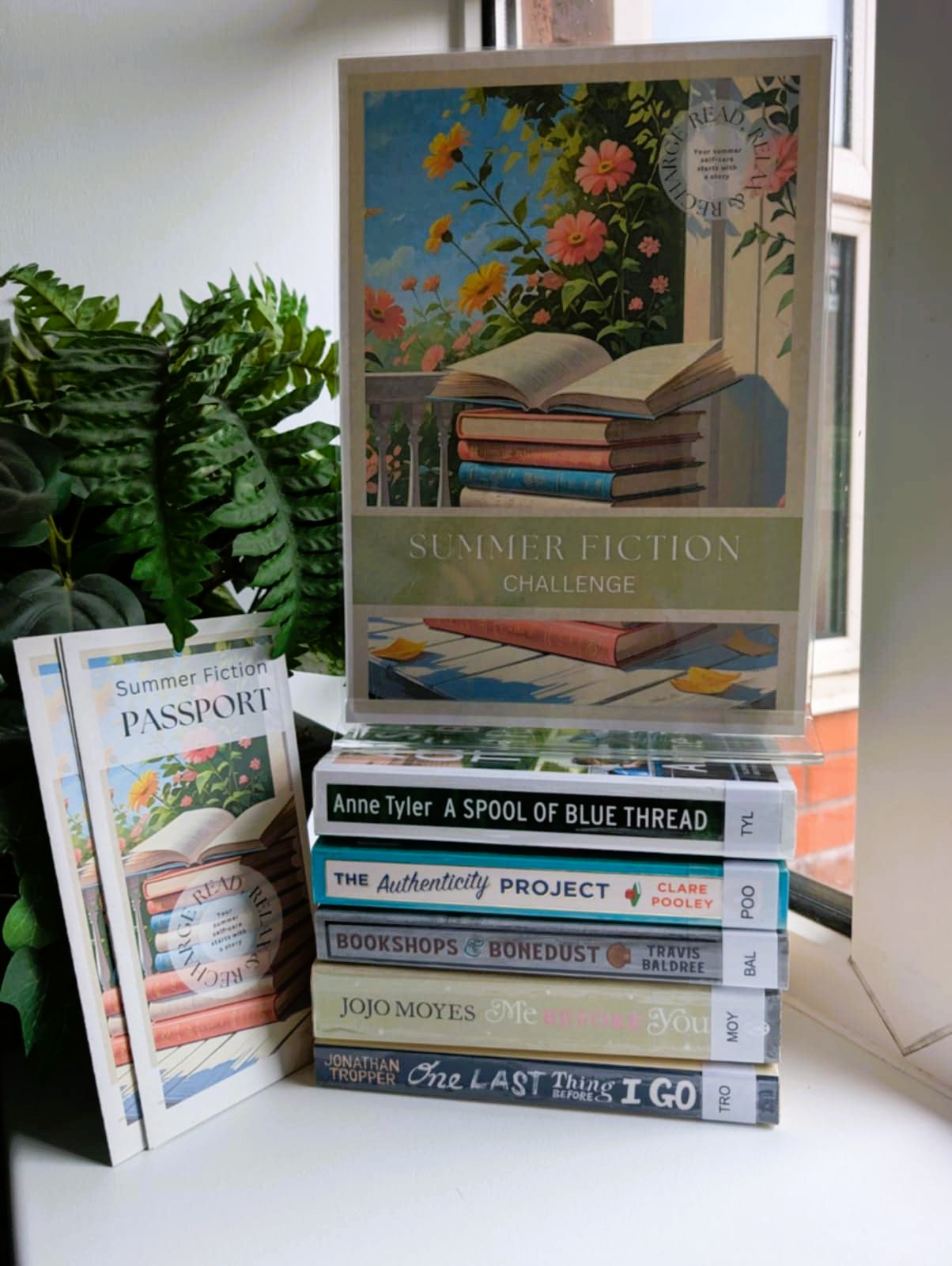 Image of a plant in the background, with some leaflets leaning against it. On the right is a pile of books and on top of the books is a poster advertising the Summer Reading Challenge